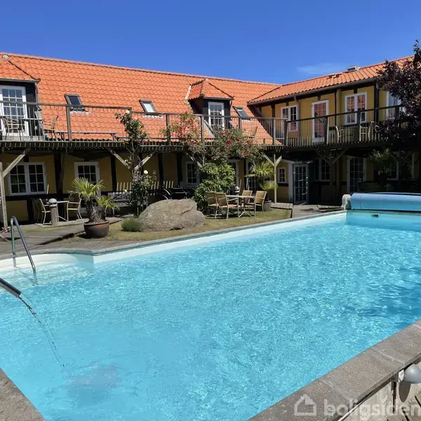 En stor udendørs swimmingpool med klart, blåt vand står centralt i en gårdhave omgivet af en gulmalet bygning med rødt tegltag, træterrasser, planter og havemøbler.