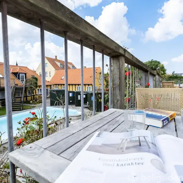 En træterrasse med et bord, hvor der ligger magasiner og bøger. I baggrunden ses en pool omgivet af huse med røde tage. Magasinet har teksten "Kunsthåndværk med karakter".