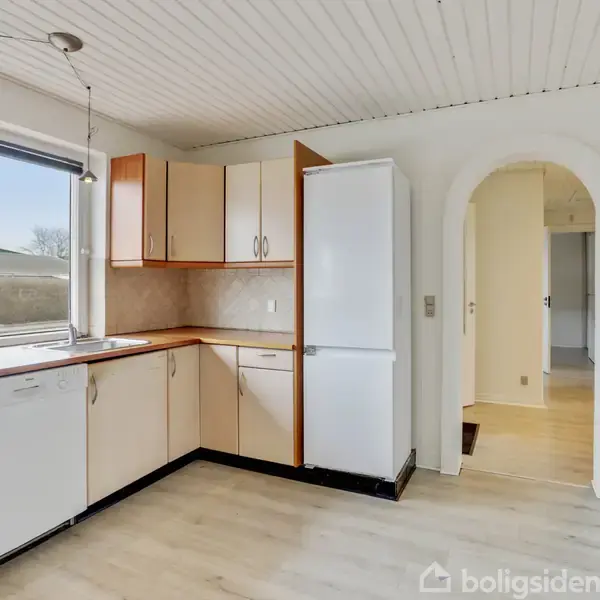 Et lyst, tomt rum med trægulv og hvidmalede vægge. En bueåbning fører til et tilstødende rum. Indbyggede loftspots tilbyder belysning.