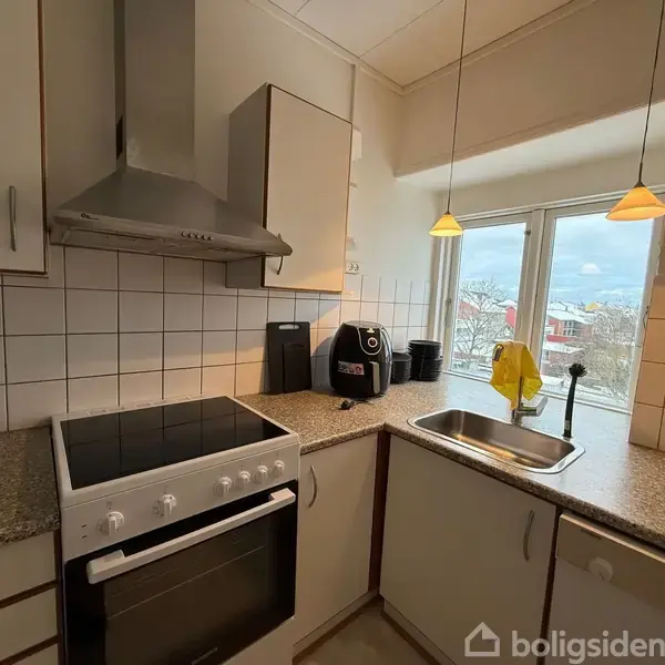 Komfur står i køkkenhjørnet ved siden af en luftudsugningshætte. Bordpladen er grå, og der står en kaffemaskine og en saftpresser ved siden af vasken. Vindue med udsigt.