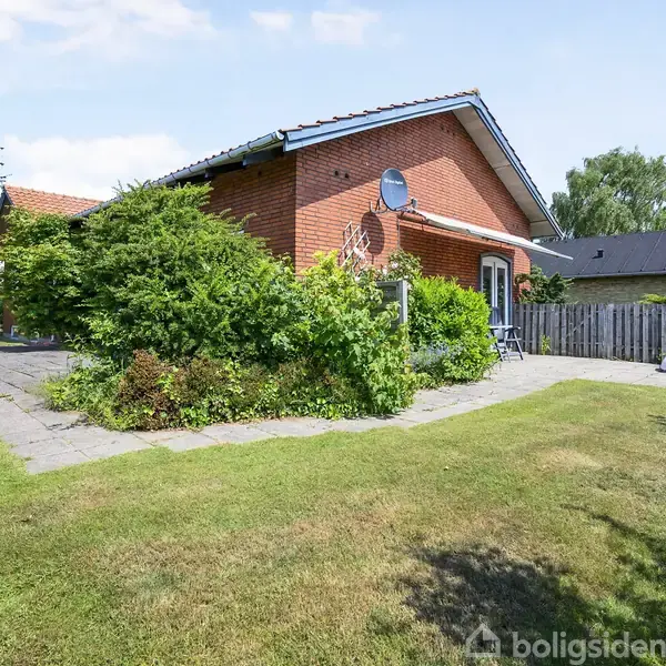 Rødt murstenshus omgivet af frodige buske i en velplejet have, med flisegang og hvid stakit til højre. Solsenge står ved stakittet.