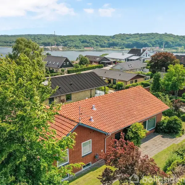 Et rødt murstenshus med rødt tegltag omgivet af frodige grønne træer og velplejede haver. I baggrunden ses flere huse og en bred søflade under en klar blå himmel.