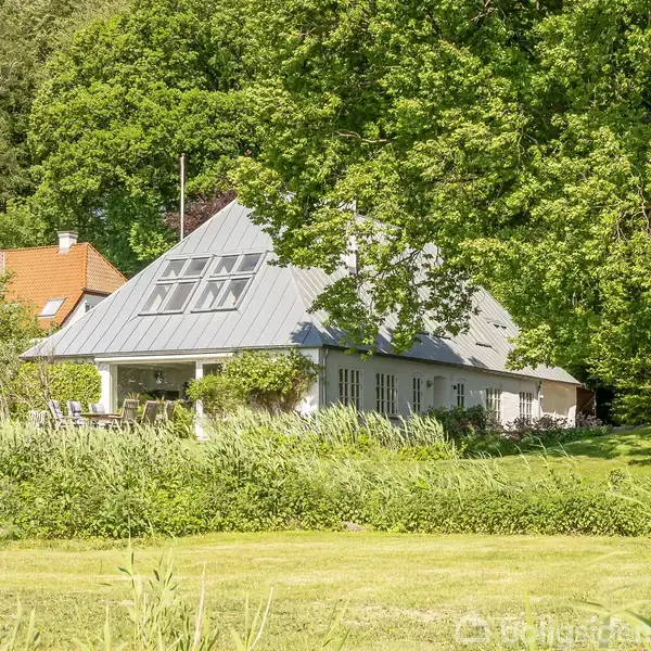 Et hvidt hus med skråt tag står blandt frodige træer og buske. Sollys skinner ned gennem grenene, og en græsplæne strækker sig foran huset.
