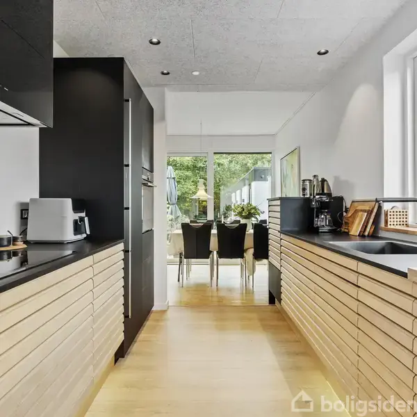 Et moderne køkken med træpaneler og sorte bordplader. Der er diverse køkkenapparater og en knivblok på bordet. I baggrunden ses et spisebord foran store vinduer med udsigt til haven.