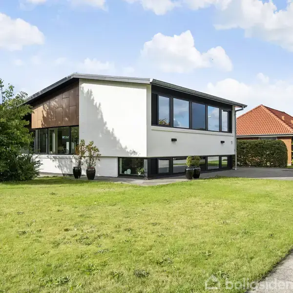 Huset står stille i en grøn have med plæne og buske. Det har en moderne facade med store vinduer. En carport med rødt tag ses i baggrunden. Skiltet viser "TIL SALG".