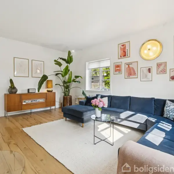 Stue med blå hjørnesofa står på et lyst tæppe; sofabord med blomster, brændeovn til venstre, og en skænk med planter og kunst på væggen skaber en hyggelig atmosfære.