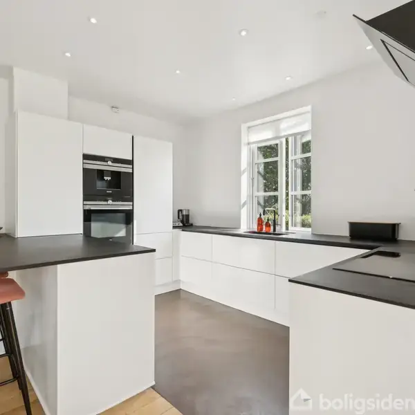 Et moderne køkken rummer en sort bordplade på en L-formet køkkenø med indbygget ovn og skabe. Lyse vægge og et vindue baggrundsbelyser rummet med naturligt lys.