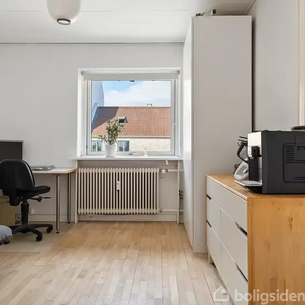 En kontorstol står foran et skrivebord med computer i et lyst rum. Til højre er en kommode med en printer ovenpå. Vindue viser udsigt til hustage.