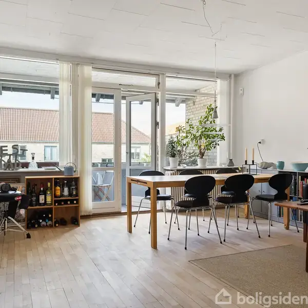 Stue med spisebord og sorte stole står på lyst trægulv. En stor plante er midtpunktet på bordet. En balkon kan ses gennem de store vinduer.