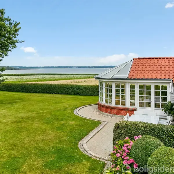 Et hus med rødt tegltag og glaseret veranda står ved en velplejet græsplæne. I baggrunden ses en sø eller fjord samt en hæk langs kanten af plænen.