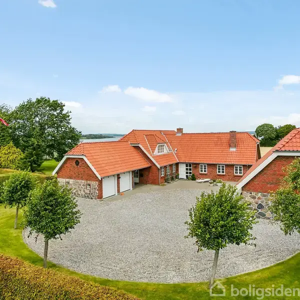 Et rødt murstenshus med rødt tegltag står omgivet af en grusbelagt gårdsplads. Flagstang med flag til venstre, grønne græsplæner og træer i baggrunden under en blå himmel.