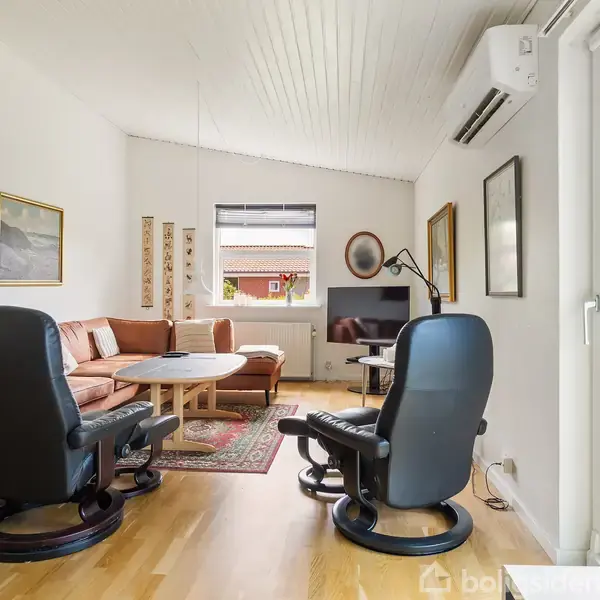 To lænestole vender mod en brun hjørnesofa med sofabord i stuen. Vægge har kunst, og et tæppe ligger på trægulvet. Udenfor ses en terrasse med borde og stole.