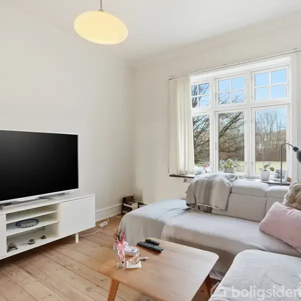 TV står på et hvidt møbel, fjernbetjening ligger på et sofabord, og en sofa med puder står ved vinduet. Lyse vægge og trægulv i en stue.