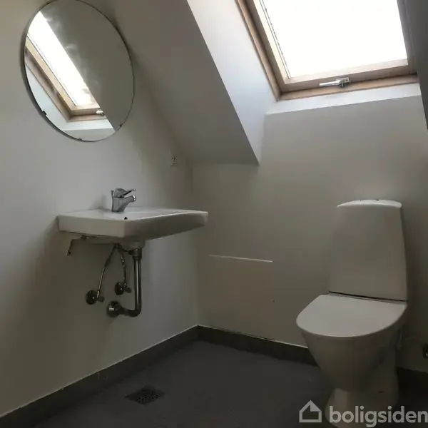 Håndvask og toilet står i et badeværelse med skråloft og ovenlysvindue. En rund spejl hænger over vasken. Skinnende armatur og rør er synlige under vasken.