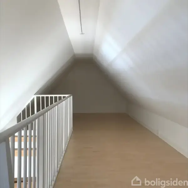 Loft med skrå vægge fører til en spids ende, omgivet af lyse trægulve og hvide vægge. Et hvidt gelænder beskytter trappen til venstre.