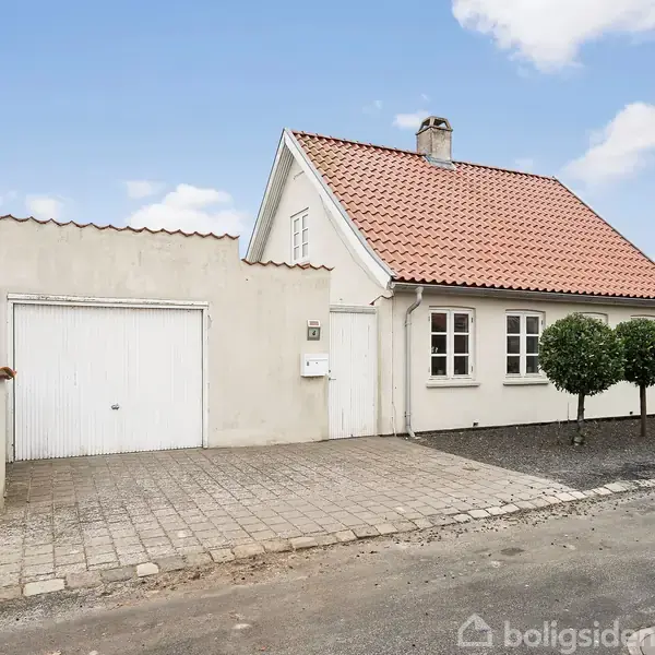 Et hvidt hus med rødt tegltag står ved siden af en garage med en lukket hvid port. Foran huset er flisebelægning og små kugletrimmede træer.