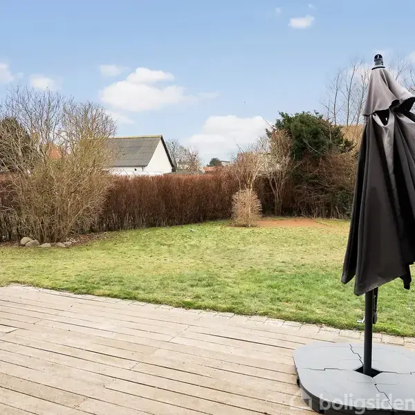 En lukket parasol står på en terrasse af træ. Græsplænen omgiver terrassen, og buske samt huse ses i baggrunden under en blå himmel.