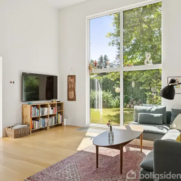 Stue med sofa og bord ved stor trærammevindue, åbner mod en grøn have. Væghængt TV og bogreol til venstre. Lyst trægulv og tæppe fuldender rummet.