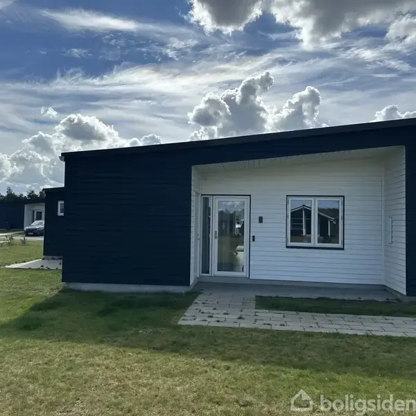 Et moderne, én-etagers hus med mørk facade og hvid indgang, stands på en grøn græsplæne under en skyet himmel. Adgangsstien er flisebelagt, omgivet af åbne områder og lignende bygninger.