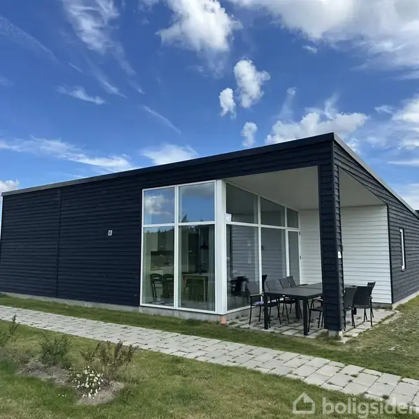 En moderne bungalow har mørke træpaneler og store glaspartier. Foran står et bord med stole på en flisebelagt terrasse. Omkringliggende græsareal og blåt skyet himmel ses i baggrunden.