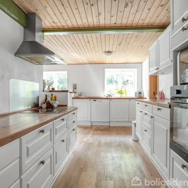 Et moderne køkken med hvide skabe og en træbordplade. Rum udsmykket med planter og køkkenudstyr. Trægulv og træbeklædt loft skaber en hyggelig atmosfære. Mål: 0-525-1000.
