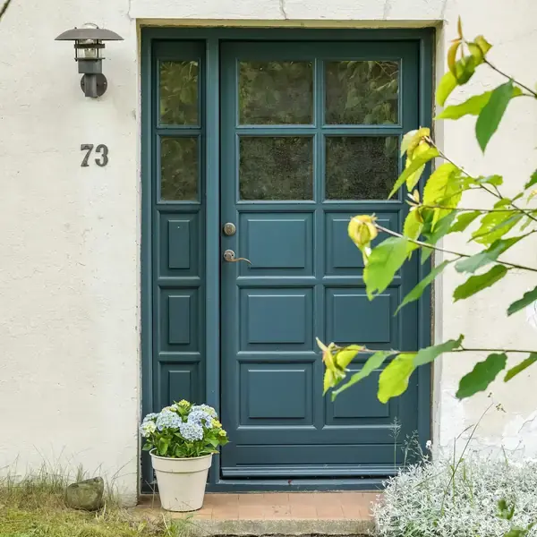 Blågrøn hoveddør med glasfelter, flankeret af husnummeret 73, omgivet af en hvid mur. Foran døren står en potteplante med lyseblomstrede hortensiaer. I højre forgrund ses grønne grene.