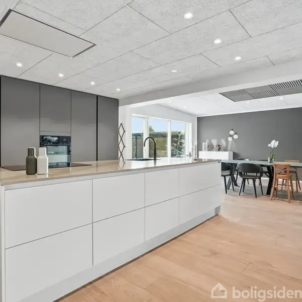 Et moderne køkken med en stor hvid køkkenø, hvor der står to dekorative flasker. I baggrunden ses et spisebord omgivet af stole i en lys stue med trægulv.