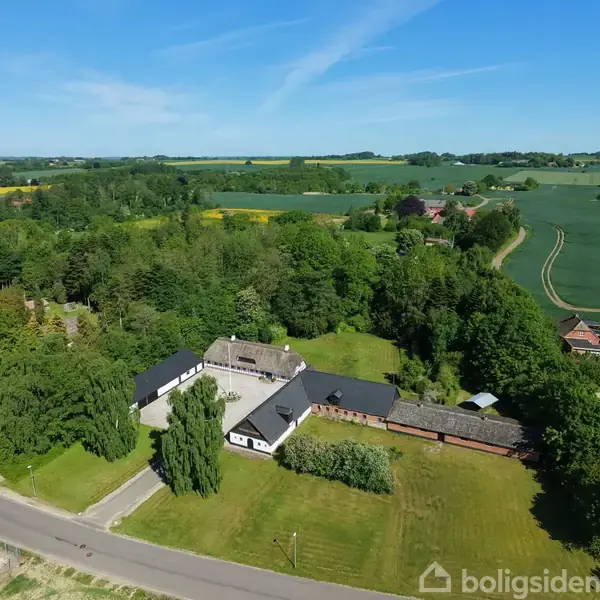 En gård med hvidmalede bygninger og stråtag står omgivet af grønne marker og træer. En landevej strækker sig langs grunden i et landligt område med spredte huse.