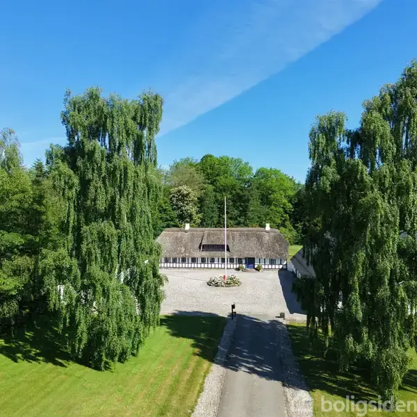 Et bindingsværkshus med stråtag står i midten omgivet af høje træer. En grusvej fører op til huset gennem en velplejet have under en klar blå himmel.