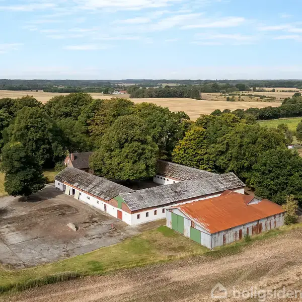 En landbrugsbygning omgivet af skov udføres ved at stå stille blandt marker. Bygningen har et rødt tag og er placeret i et åbent, bakketagende landskab under en blå himmel.