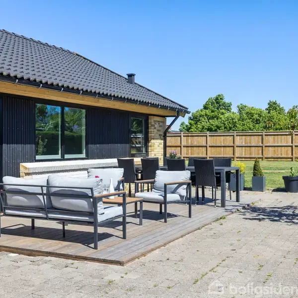 Terrasse med havemøbler, herunder en sofa og stole, står foran et moderne hus med sorte træfacader og store vinduer. En træterrasse omgivet af fliser og grønne buske i baggrunden.
