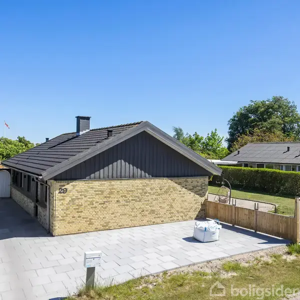 En gul murstensbungalow med sort tag står stille på en flisebelagt indkørsel. Ved siden af huset er en græsplæne og træhegn. Husnummeret 23 er synligt.