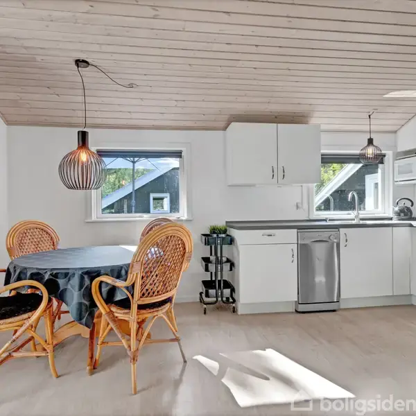 Et lyst køkken med hvide skabe og stålapparater. Et rundt bord med fire kurvestole står ved siden af to vinduer. En hængelampe hænger over bordet.