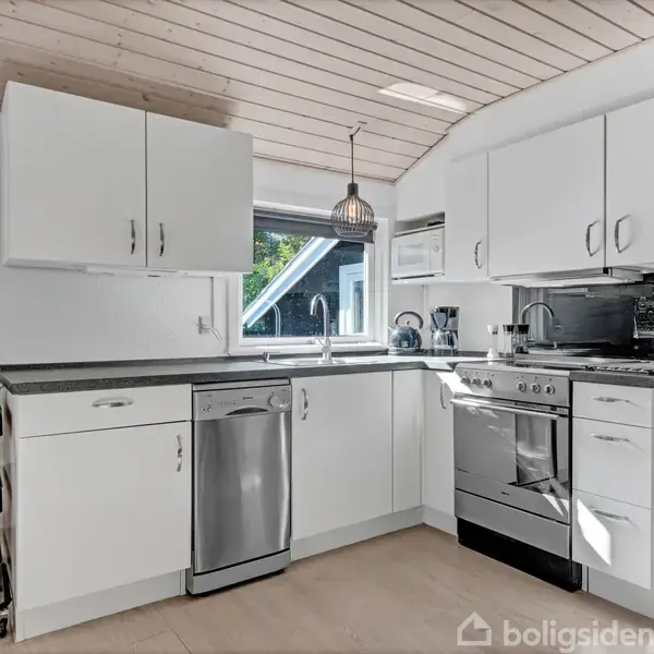 Et stilrent, hvidt køkken med sorte bordplader, indeholder en opvaskemaskine, komfur og vask. Køkkenet har et skråt træloft og vinduer, der giver naturligt lys. En sort hylde står til venstre.