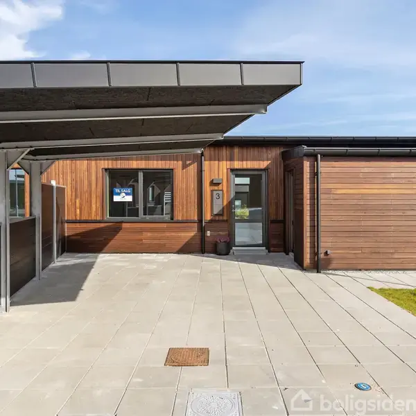 Carport foran et moderne træhus med fladt tag; belægning af lyse fliser i forgrunden; plæne til højre side; huset har store vinduer og en indgangsdør; del af et boligområde.