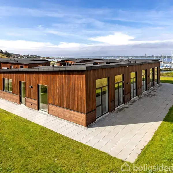 Bygningen med fladt tag og træfacade står stille omkranset af grøn græsplæne og flisebelægning, beliggende tæt ved en bådehavn under en delvist skyfri himmel.