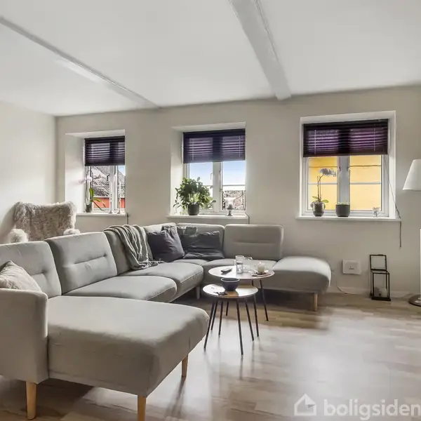 Grå sofa står i lys stue med tre vinduer med persienner. Stuebord med dekorationer foran sofaen. Hvidt skab og lampe i hjørnet. Trægulv og lys vægfarve.