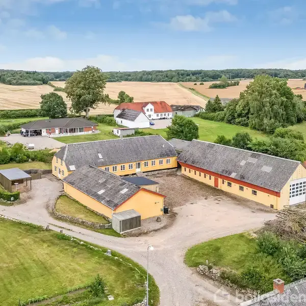 Landbrugsejendom med gule bygninger omkring en central gårdsplads. Omgivet af marker og træer i et landligt landskab. Flere mindre bygninger og veje forbinder ejendommene.
