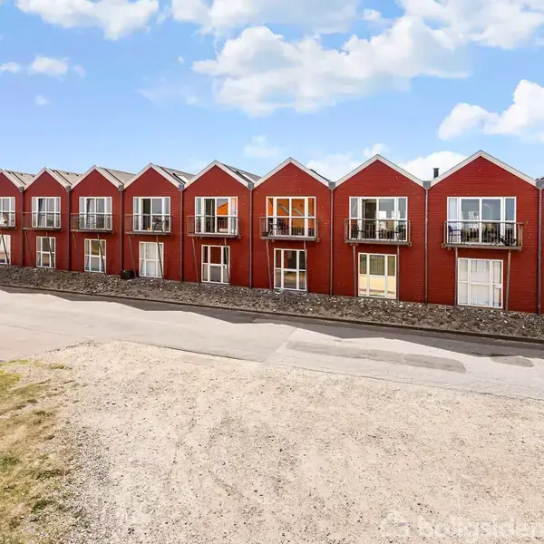 Række af røde byhuse med altaner står stille langs en vej. Foran er en grusvej og græsareal. Et skilt med teksten "P forbudt" ses på venstre bygning.
