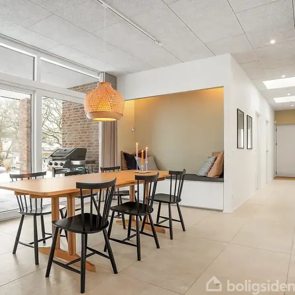 Et lyst køkken-alrum med et træbord omgivet af sorte stole. Store vinduer i baggrunden giver udsigt til en terrasse med havemøbler og en grill. Plante i forgrunden til højre.