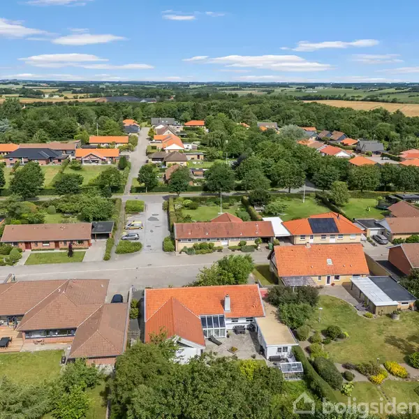 En række huse med røde tage står stille i et grønt villakvarter, omgivet af velplejede haver og træer. Biler er parkeret på vejen mellem husene under en blå himmel.