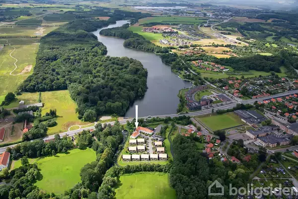 Bebyggelse er markeret med en hvid pil og omkranset af tæt skov. Området ligger nær en snoet flod, med marker og huse omkring. Billedet viser et bredt landskab.