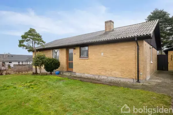 Et gult murstenshus står stille i en lille have med træer og græsplæne. Huset har en brun dør og vinduer samt et gråt tag.