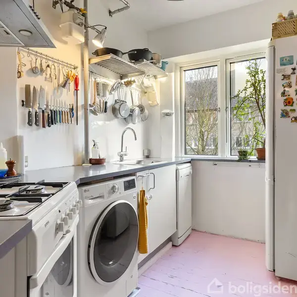 Et funktionelt køkken med komfur og vaskemaskine ved siden af hinanden. Køkkenredskaber hænger organiseret på væggen. Store vinduer giver naturligt lys. Køleskabet har forskellige magneter.