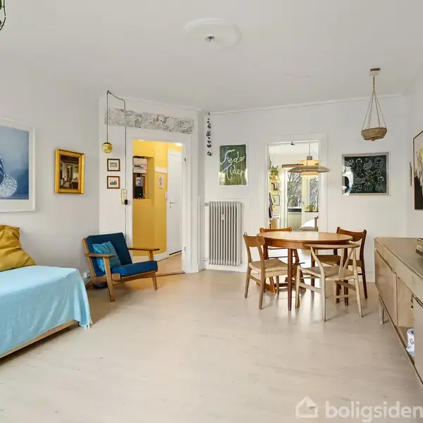 En stue med en blå sofa og et rundt spisebord med fire stole. Indretningen har træmøbler og mange billeder på væggene. Lyse vægge og et vindue til venstre giver rummet lys.