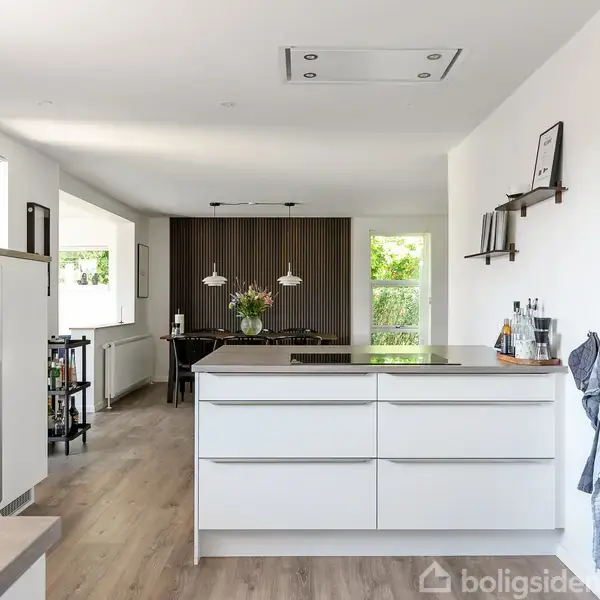 Et moderne køkken med hvide skabslåger og en stor ø i midten, omkranset af lys træparket. I baggrunden ses spiseplads med bordlamper og store vinduer, der giver udsigt til naturen.