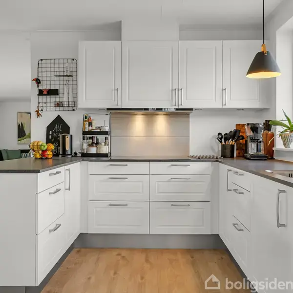 Moderne køkken med hvide skabe omkranser et hjørne. En frugtskål og køkkenredskaber står på bordet. En metalkogeplade er nedenfor en emhætte, og et vindue til højre lader lys ind.