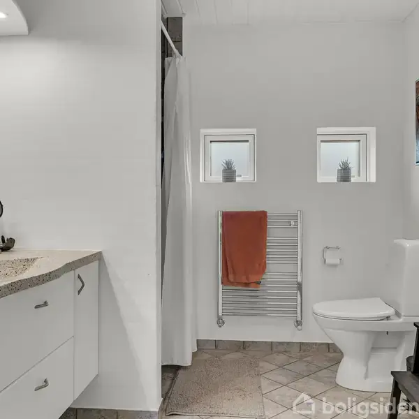 Badeværelse med hvidt interiør; en stenbordplade med vask, ved siden af et toilet og brusebad. To små vinduer og en opvarmet håndklædetørrer med et orange håndklæde.