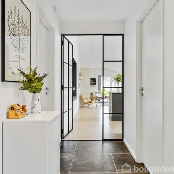 En sort metalrammet glasdør åbner ind til en lys stue med moderne møbler. På skænken står en vase med blomster og en dekorativ kalender, der viser datoen "01.08".