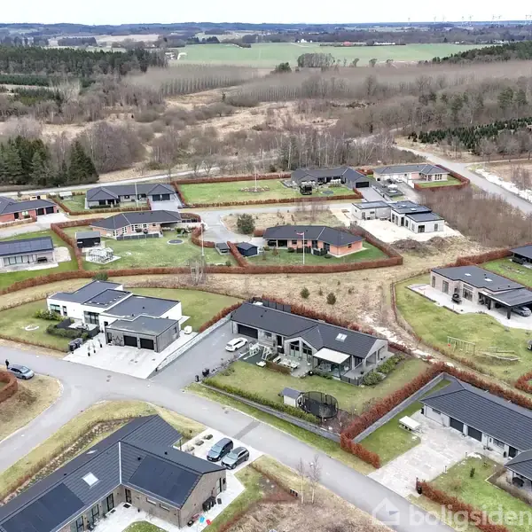 Boliger ligger i et moderne kvarter med huse spredt langs krumme veje. Husene er omgivet af græsområder og hække, med landlige omgivelser af skov og marker i baggrunden.